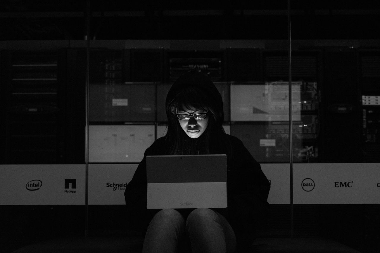 our-story-01 A woman in a dark room uses a laptop, illuminated by its screen, with server equipment in the background.